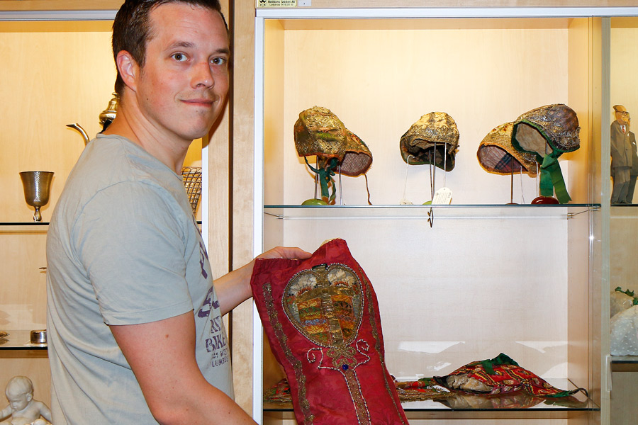 Joakim Karlsson håller fram en doppåse från 1700-talets första hälft. I bakgrunden syns resten av samlingen av dopmössor och doppåsar från 1600-1800-tal.